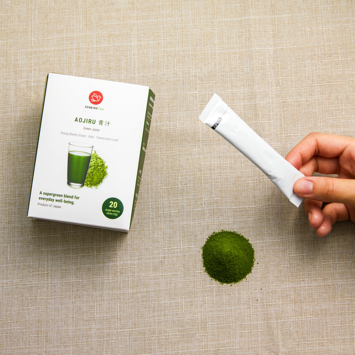Hand holding an Aojiru single-serving stick beside Senbird Japanese green juice powder and its box on beige linen