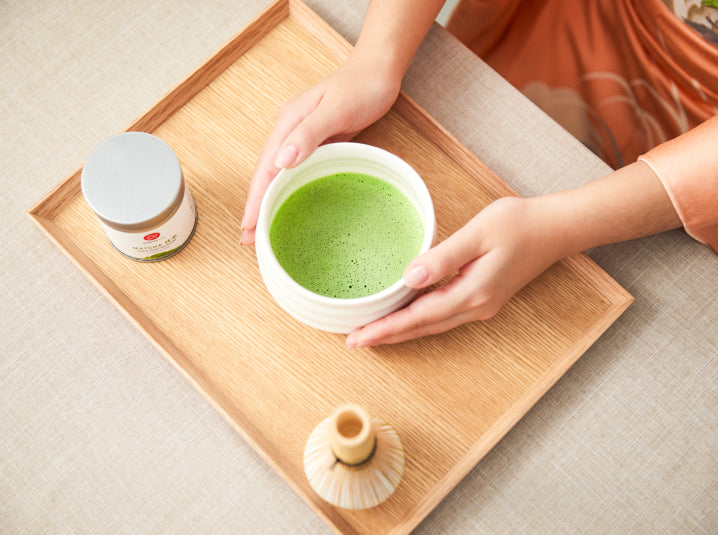 hand holding brewed matcha in a cup with matcha tea tin and bamboo whisk
