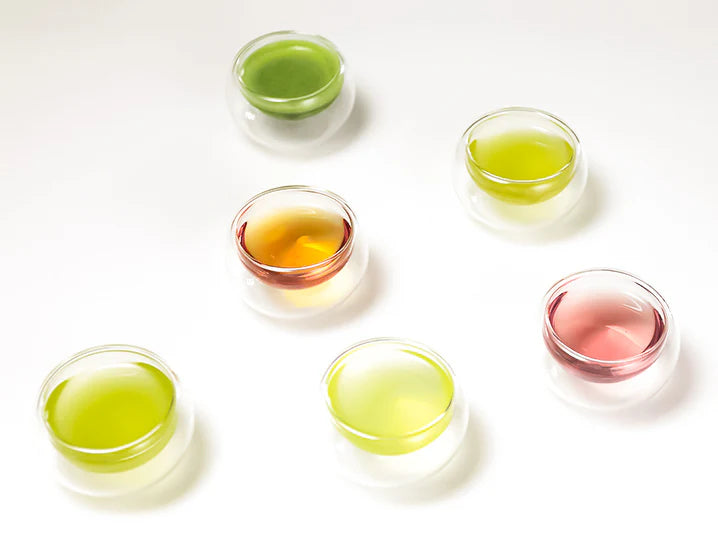 Overhead view of six double-walled glass cups showing the beautiful color range of Japanese teas from vibrant green matcha to pale yellow-green sencha to golden amber hojicha