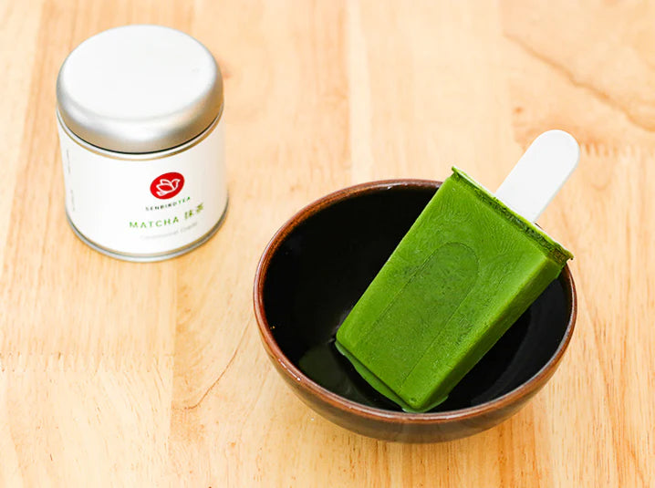 Senbird Tea ceremonial grade matcha tin beside a vibrant green matcha popsicle resting in a dark ceramic bowl on a warm wooden surface