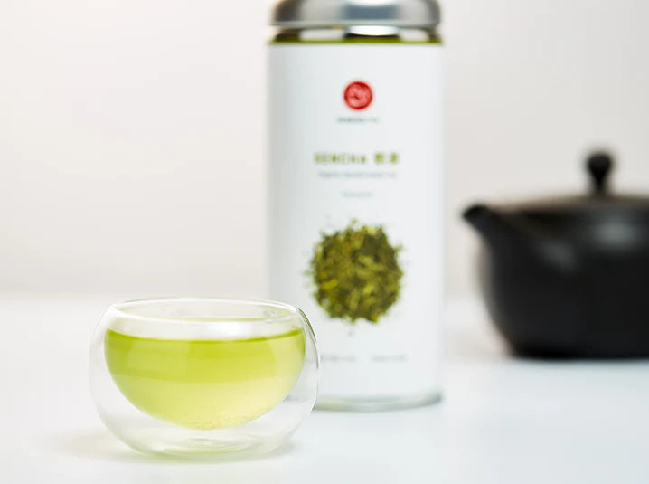 Brewed sencha Japanese green tea in a double-walled glass cup with a Senbird Tea tin and kyusu teapot in the background