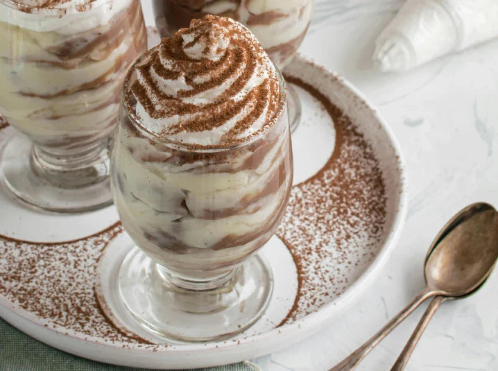 Slice of hojicha tiramisu on a gray plate with a gold spoon alongside two individual tiramisu ramekins dusted with hojicha powder on a white surface