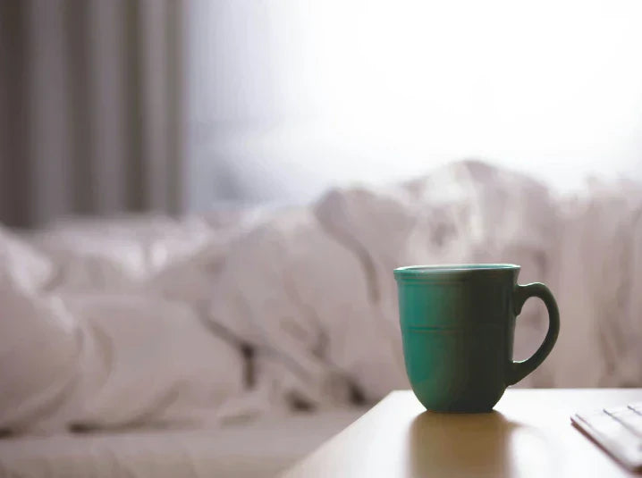 A green mug of warm tea resting on a bedside table next to soft white bed linens in muted, dreamy tones