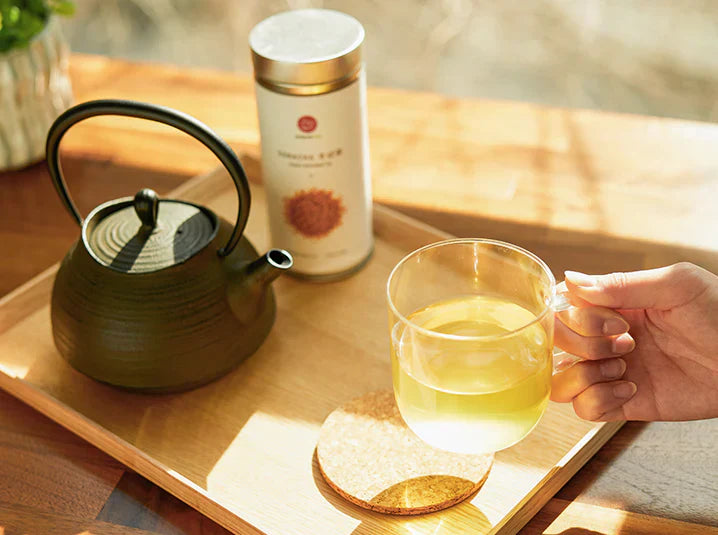 A hand lifts a clear mug of tea. A black kettle and tea tin sit on a wooden tray.
