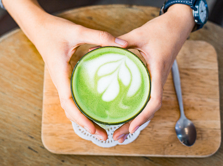 hot matcha green tea latte with a nice heart shaped latte art in a glass cup on top of a dark wooden coaster