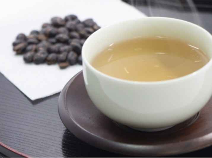 On a wooden surface, there is a pile of kuromame, or black soybeans on a white napkin to the left. On the right side, there is kuromamecha, or black soybean tea, in a white teacup on top of a wooden saucer.