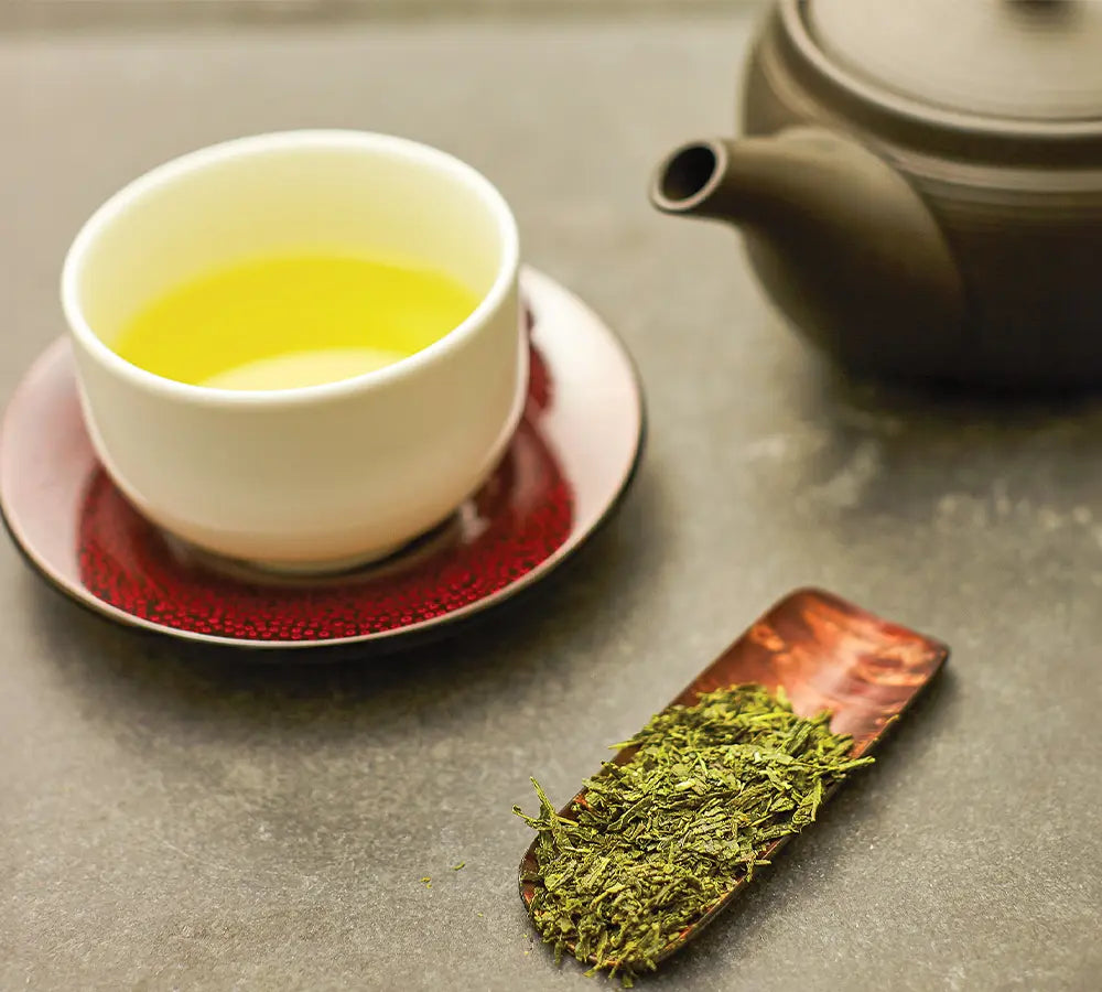 Japanese green tea served in a white cup with loose tea leaves on a wooden scoop and a dark teapot