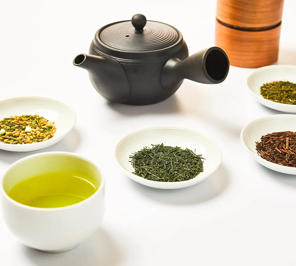 Black teapot with a cup of Japanese green tea and assorted loose leaf teas in white tasting dishes
