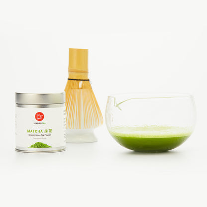 clear glass matcha bowl with whisked matche beside matcha tin and matcha whisk and stand