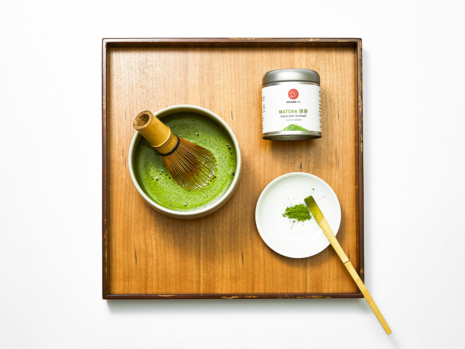 matcha tea powder tin, bamboo whisk stirring tea in a match bowl & bamboo scoop with tea powder in a plate